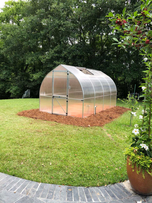 Backyard installation of Exaco Hoklartherm Riga 4 greenhouse placed on a mulched base, framed by green lawn and surrounded by trees and plants.