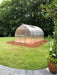 Backyard installation of Exaco Hoklartherm Riga 4 greenhouse placed on a mulched base, framed by green lawn and surrounded by trees and plants.