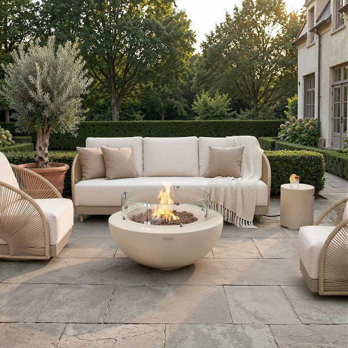 Wide golden-hour lifestyle photo of the Elementi Lunar Bowl Fire Table burning on a stone patio surrounded by wicker lounge chairs, a beige sofa, a side table holding a drink, a potted olive tree, manicured hedges, and a European-style home facade at sunset.