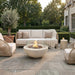 Wide golden-hour lifestyle photo of the Elementi Lunar Bowl Fire Table burning on a stone patio surrounded by wicker lounge chairs, a beige sofa, a side table holding a drink, a potted olive tree, manicured hedges, and a European-style home facade at sunset.