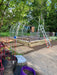 Backyard installation of Riga greenhouse frame with aluminum arches, ladders, and wood base, surrounded by garden plants and starter vegetable beds.