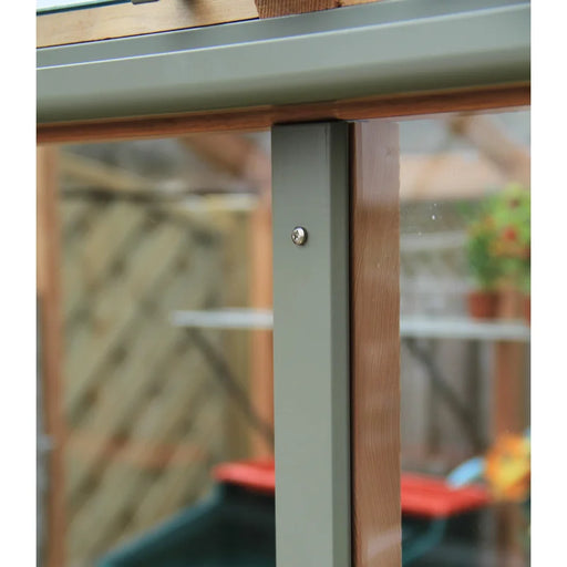 Close-up of a corner joint in the Alton Cedar Structure Fusion Greenhouse by Backyard Oasis, highlighting secure glazing with a screw fastening the frame. Blurred garden items and greenhouse accessories appear in the background.