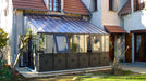 A black aluminum lean-to greenhouse with decorative accents, glass panels, and roof vents, attached to a house with a tiled roof.