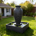 Black amphora fountain with square basin, gentle water flow and floating plants in contemporary backyard landscape.