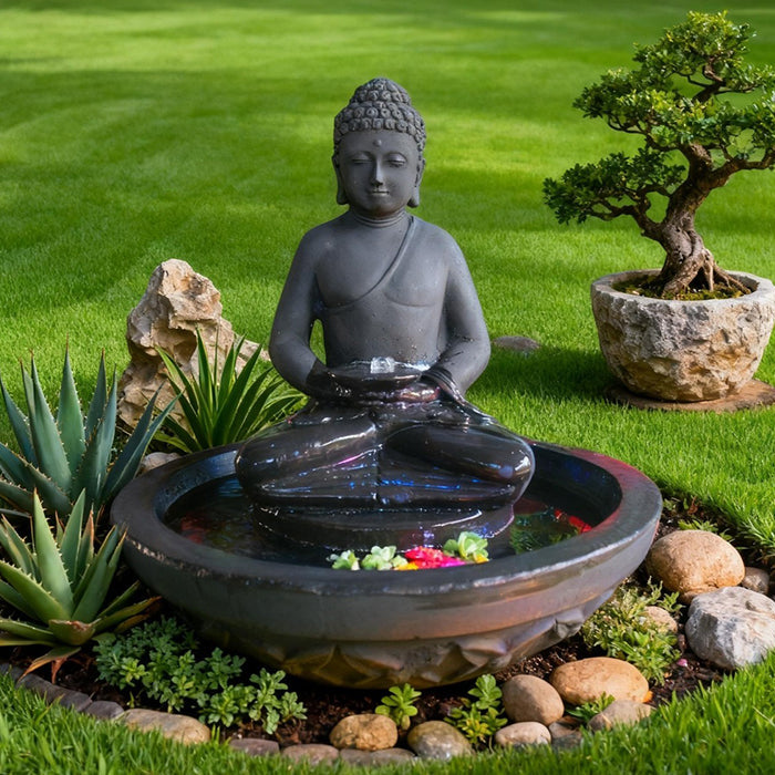 Black Buddha outdoor water fountain with bowl basin and subtle LED light, surrounded by green lawn, stones, and plants.