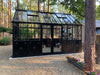 Front entry view of the Retro Royal Victorian VI Greenhouse in black with ornate decorative lower panels, double doors, and glass roof set in a wooded garden.