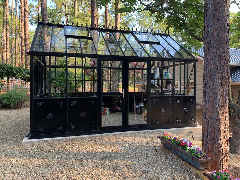 Front entry view of the Retro Royal Victorian VI Greenhouse in black with ornate decorative lower panels, double doors, and glass roof set in a wooded garden.