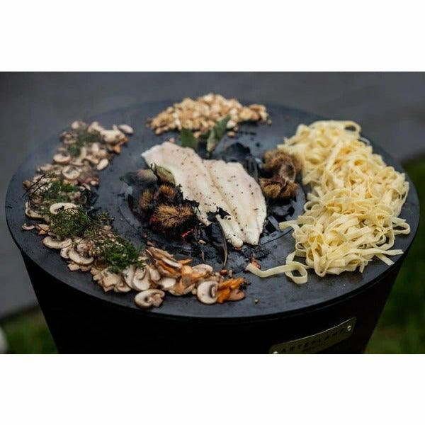 A culinary spread including mushrooms, sliced meat, and noodles on the Arteflame Black Label One Series 20" Grill's cooking surface, illustrating its versatile outdoor cooking capabilities.