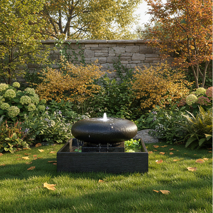 Black moon bowl outdoor fountain centered in autumn garden with stone wall backdrop and seasonal foliage.