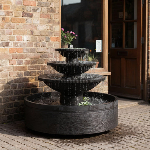 Black three tier outdoor water fountain with cascading bowls in a modern patio setting against brick wall and wooden door