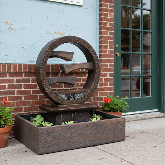 Brown resin circular water fountain with three cascading spouts and rectangular basin placed near a brick wall entryway for elegant patio décor.