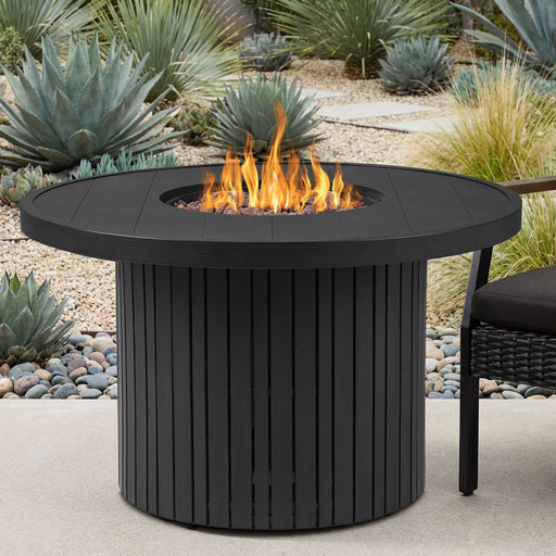 Calvin 44" Round Fire Pit Table by Real Flame lit with open flame on a concrete patio, flanked by a cushioned wicker chair amid an agave and succulent garden landscape