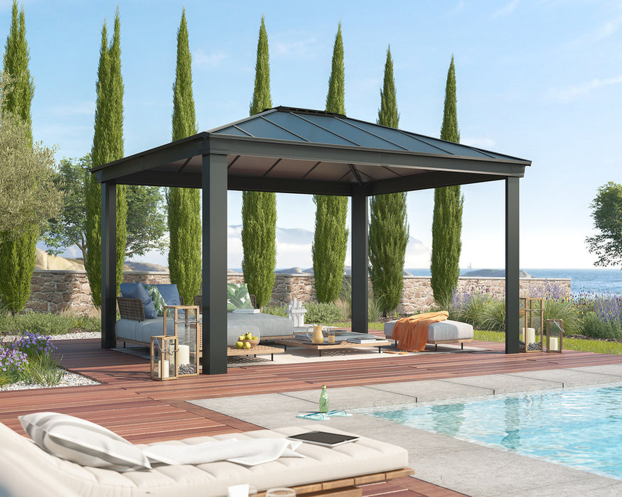 A black Canopia Dallas Gazebo with a peaked roof sits on a patio beside a pool. The patio is furnished with lounge chairs, a sofa, and a coffee table. Several tall, slender trees are planted nearby.