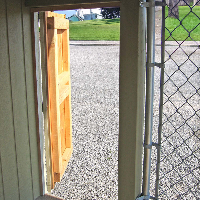 Interior view of the Cape Cod Cozy Kennel wooden door open to the outside, showing durable framing and chain-link fencing.