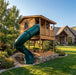 Left-angle view of the Outdoor Living Today Cozy Cabin Playhouse with tube slide and stone-lined base near a stone estate home