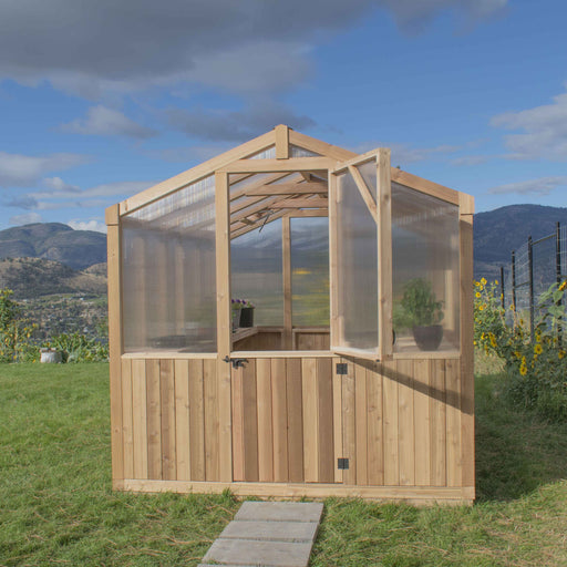 A wooden Cedar greenhouse 8x12 by Outdoor Living Today with transparent panels, open door, and potted plants inside, set against a backdrop of mountains and blue sky.