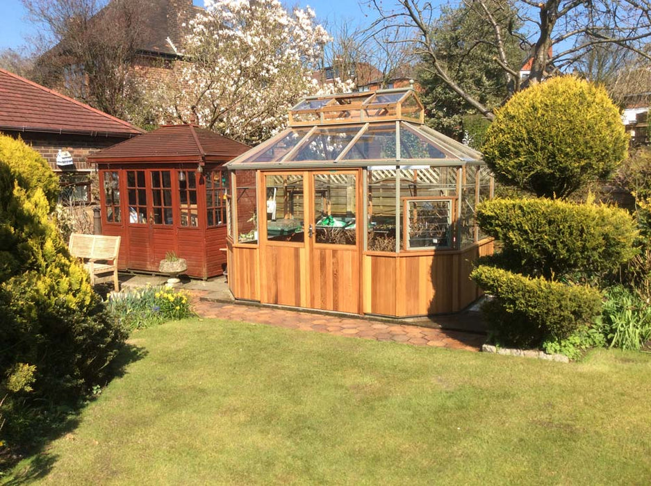 Front view of the Alton Cedar Structure Evolution Octagonal 9x11 in a garden, showcasing its elegant wooden frame and glass panels.