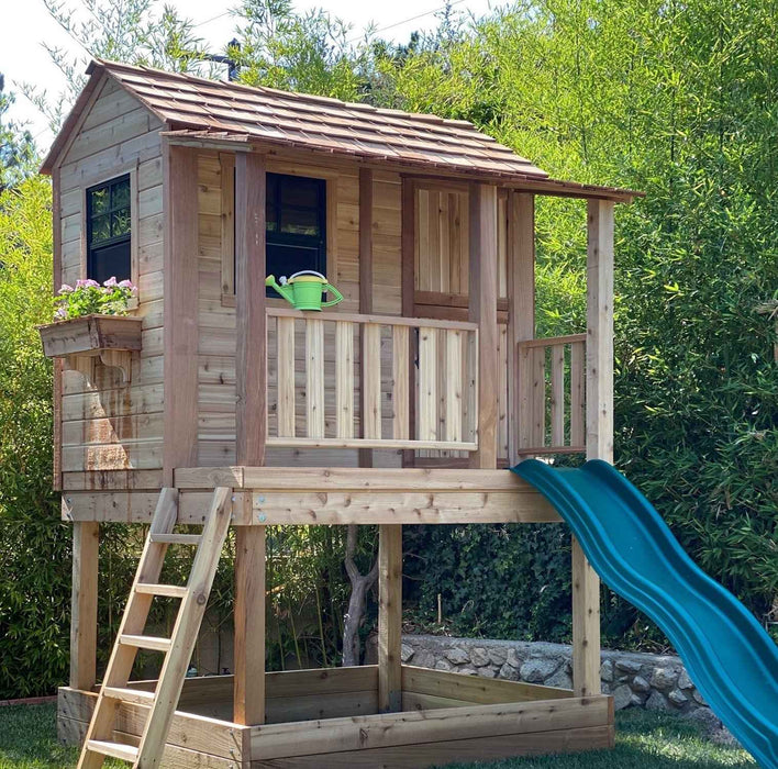 Outdoor Living Today Little Cedar Playhouse with blue wave slide surrounded by lush greenery