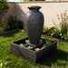 Charcoal amphora outdoor water fountain with square base, textured finish and decorative greenery in modern yard.