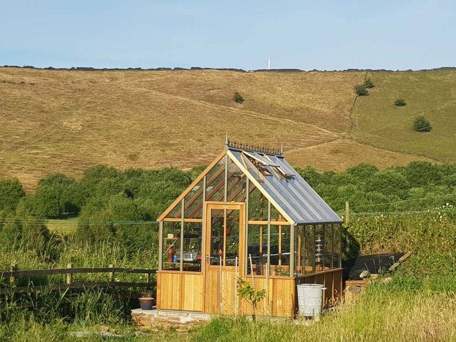 Alton Cedar Cheltenham Greenhouse placed in an open landscape with hilly terrain.