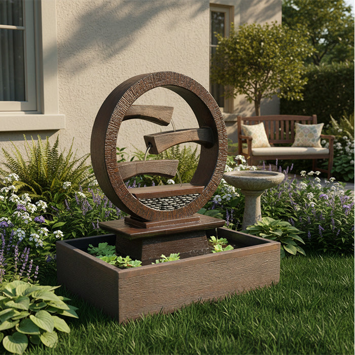 Circular tiered outdoor water fountain with textured resin frame and square basin, showcased in a lush landscaped garden with flowers and greenery.