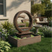 Circular tiered outdoor water fountain with textured resin frame and square basin, showcased in a lush landscaped garden with flowers and greenery.