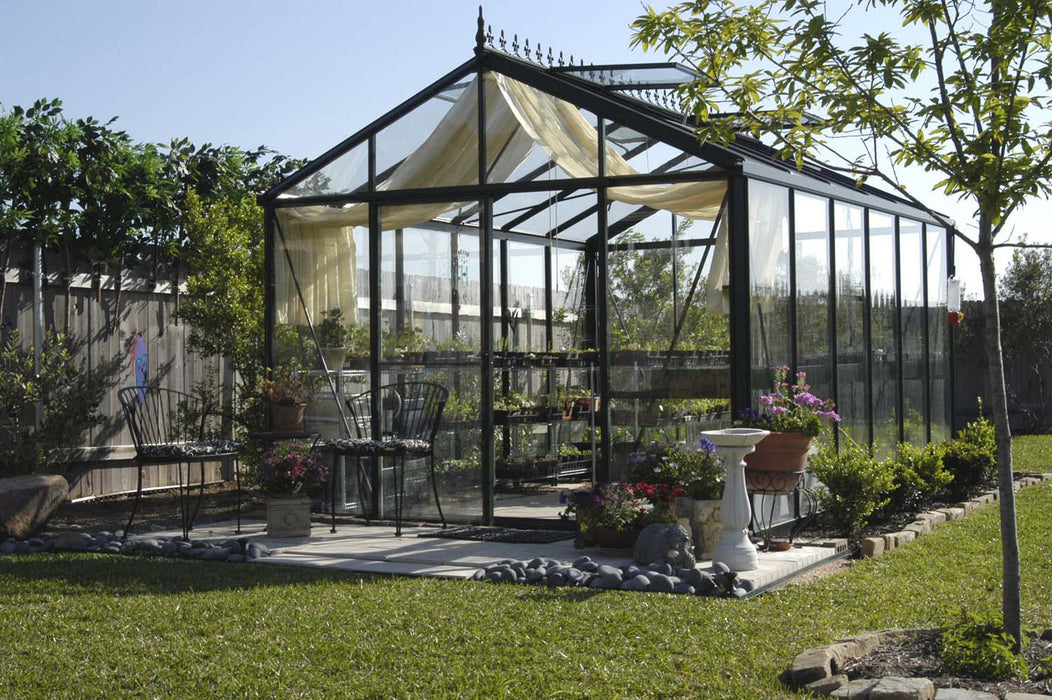 Close front view of Royal Victorian VI 34 Greenhouse with patio seating, potted plants, and decorative elements in a landscaped backyard.