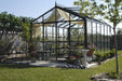 Close front view of Royal Victorian VI 34 Greenhouse with patio seating, potted plants, and decorative elements in a landscaped backyard.