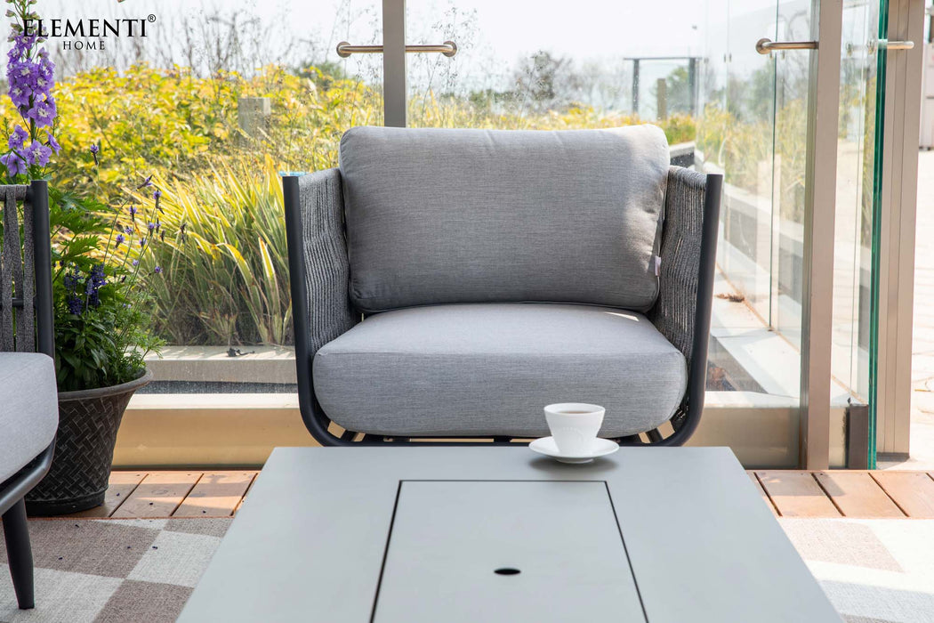 A closer view highlighting a single grey rope weave outdoor armchair with a comfortable light grey cushion. A white cup and saucer rest on a sleek grey rectangular fire table. 