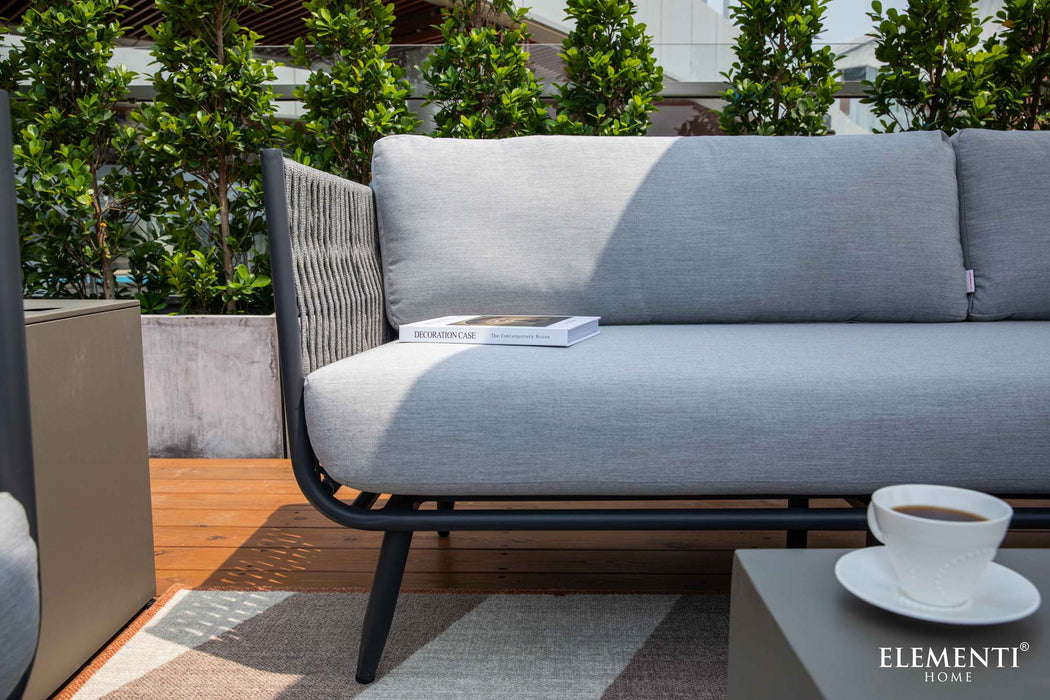A close-up of a modern grey 3 seat sofa from the Elementi Milano Collection. A book with a white cover is placed on the light grey seat cushion. 