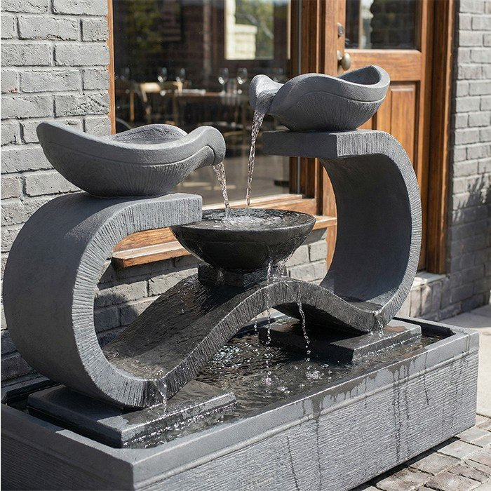 Concrete garden fountain from Exaco's Infinity line, Rust finish, set against a bamboo backdrop with flowering plants.