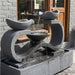 Concrete garden fountain from Exaco's Infinity line, Rust finish, set against a bamboo backdrop with flowering plants.