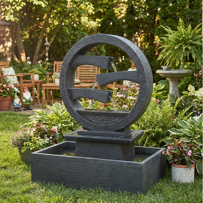 Contemporary round resin outdoor water fountain with layered waterfall design and square catch basin, displayed in a colorful backyard garden.