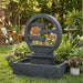 Contemporary round resin outdoor water fountain with layered waterfall design and square catch basin, displayed in a colorful backyard garden.