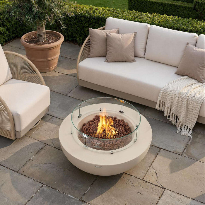 A lit cream white concrete fire pit table with a round glass wind guard sits atop a stone patio in front of a plush beige outdoor sofa with tan throw pillows and a knit blanket, flanked by a potted olive tree, creating a cozy backyard lounge seating area.