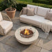 A lit cream white concrete fire pit table with a round glass wind guard sits atop a stone patio in front of a plush beige outdoor sofa with tan throw pillows and a knit blanket, flanked by a potted olive tree, creating a cozy backyard lounge seating area.