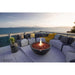 A lit dark gray concrete fire bowl glows warmly at dusk on a rooftop deck overlooking the open ocean, surrounded by a curved gray sectional sofa with mustard yellow and patterned throw pillows -- a scene styled by Elementi.