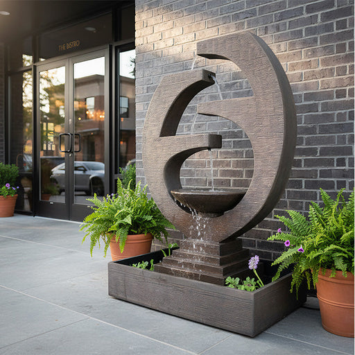 Eclipse modern outdoor water fountain with circular sculptural design placed by brick wall entryway with flowing tiered water feature.