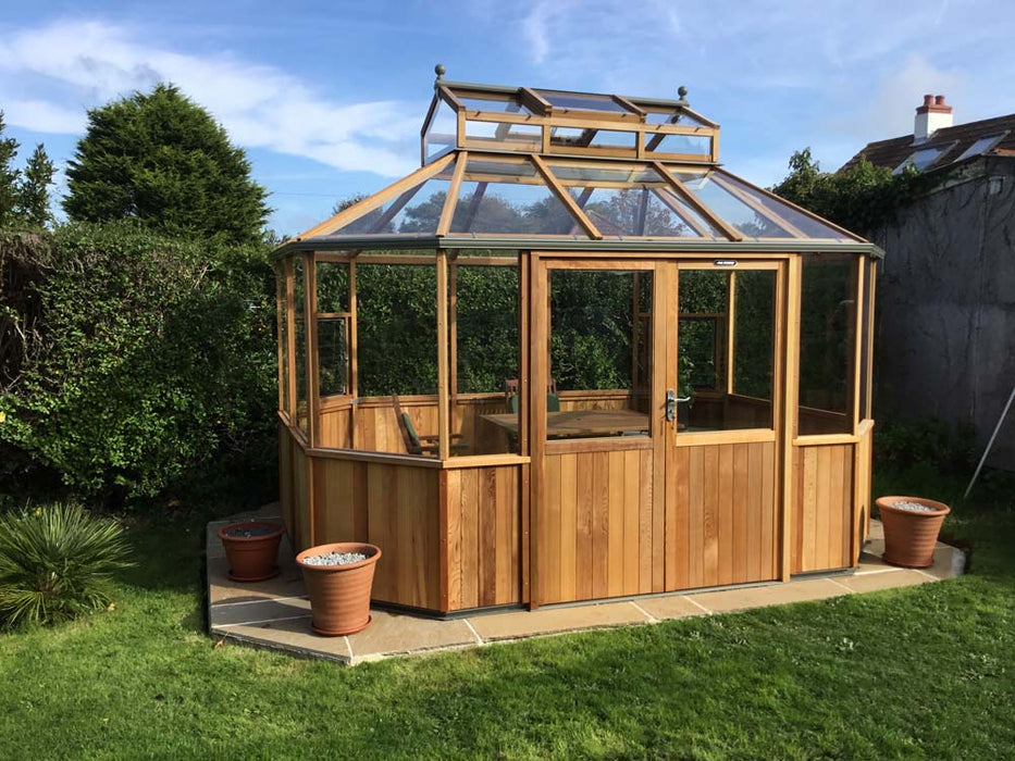 An Alton Cedar Structure Evolution Octagonal, featuring a wooden frame with glass roof and walls, is situated in a garden, embraced by lush greenery and two potted plants.