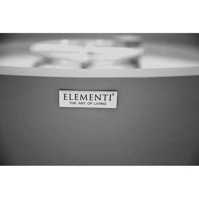 The Elementi nameplate badge reading 'The Art of Living' is mounted on the curved gray concrete exterior of the fire bowl, shown in a soft-focus black-and-white close-up with the burner ring blurred in the background above.