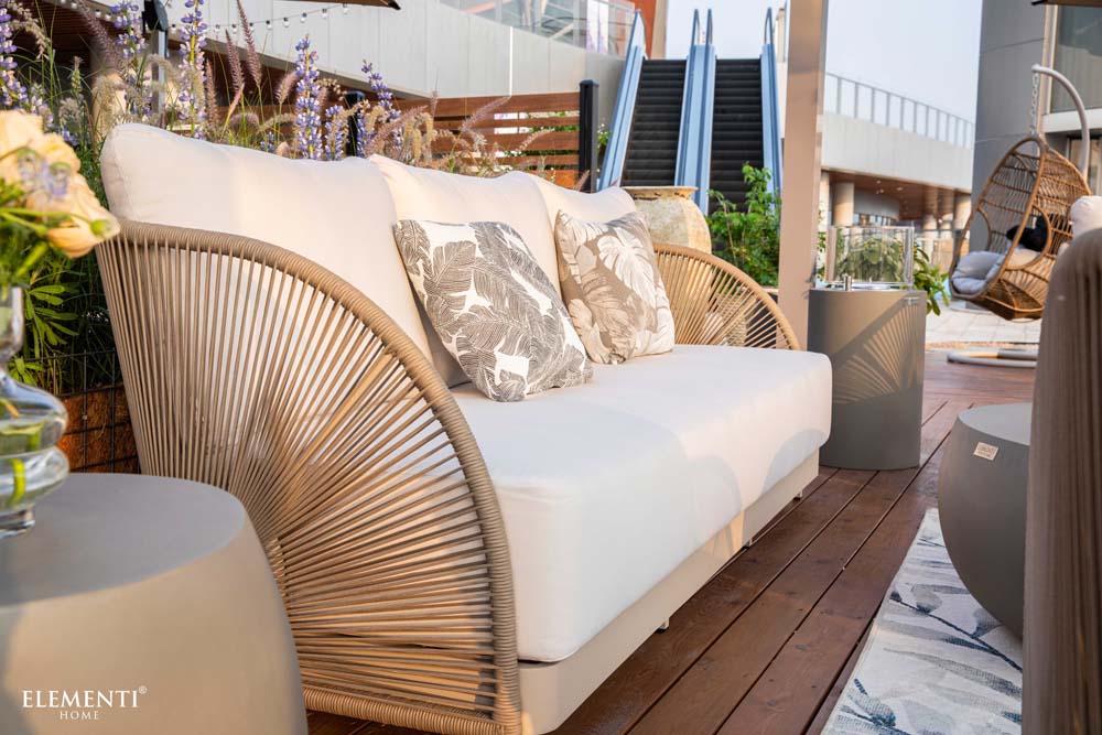 Close-up of Elementi Malibu Collection outdoor sofa featuring neutral tones and floral-patterned cushions, nestled among vibrant garden decor.