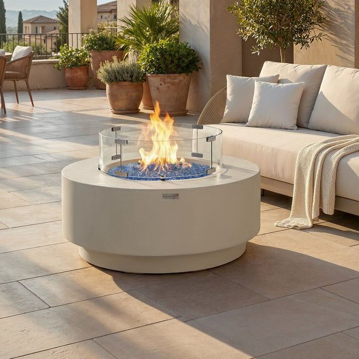Elementi Nimes fire pit table burning with glass wind guard installed, set on a stone patio beside a cushioned outdoor sofa