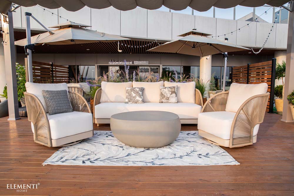 Complete view of Elementi Outdoor Furnitures Malibu Collection featuring a full patio setup with neutral chairs, sofa, and center coffee table on a deck.

