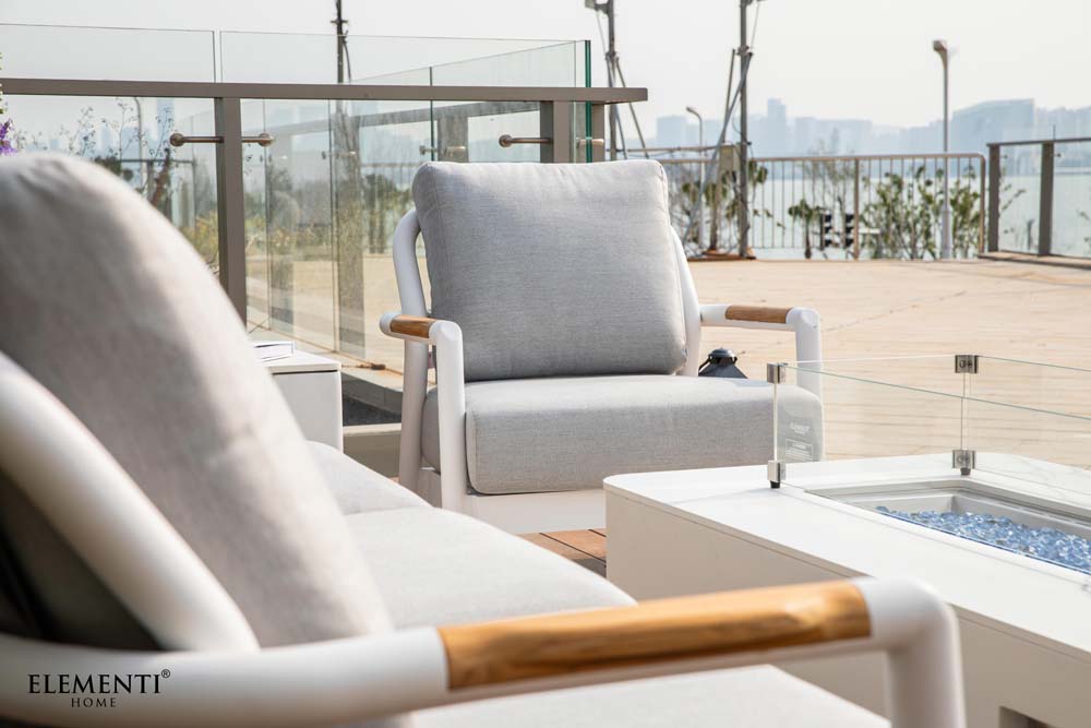 A single Elementi Santorini Collection armchair with light gray cushions and white frame, on a patio with a glass railing and city skyline.