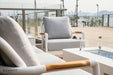 A single Elementi Santorini Collection armchair with light gray cushions and white frame, on a patio with a glass railing and city skyline.