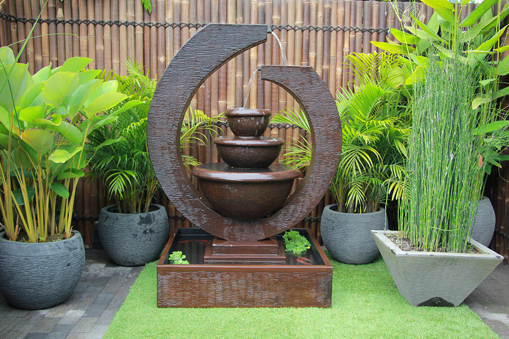 Brown Exaco Eclipse Fountain with a three-tiered water feature, bringing a modern and elegant touch to any outdoor garden space with surrounding greenery and bamboo fencing.