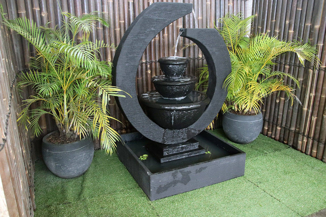 Exaco Eclipse Fountain with a three-tiered water feature in black ceramic, featuring a circular design and water cascading down into a basin with lush green plants on either side.