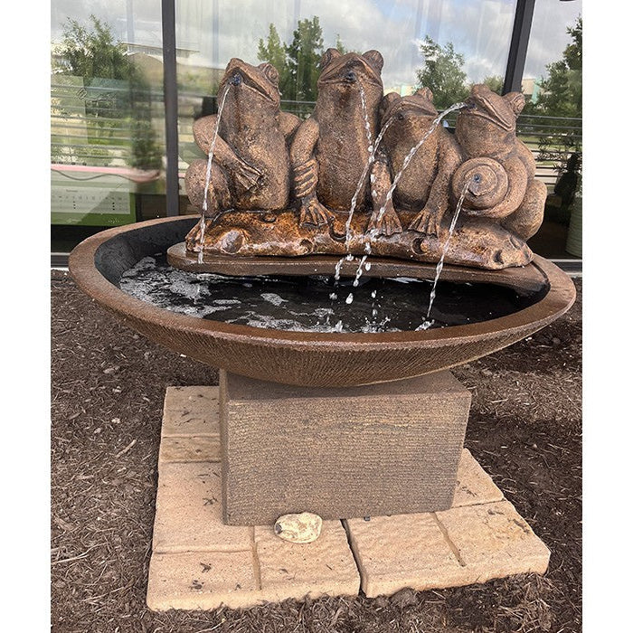 Exaco Frogs in the Pond Fountain with four frogs spouting water from their mouths, set in a large bowl-shaped basin surrounded by a bed of mulch and a modern building backdrop.