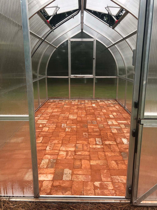 Interior of Exaco Hoklartherm Riga 4 greenhouse with clear polycarbonate walls, aluminum framing, roof vent, and rustic brick flooring installation.