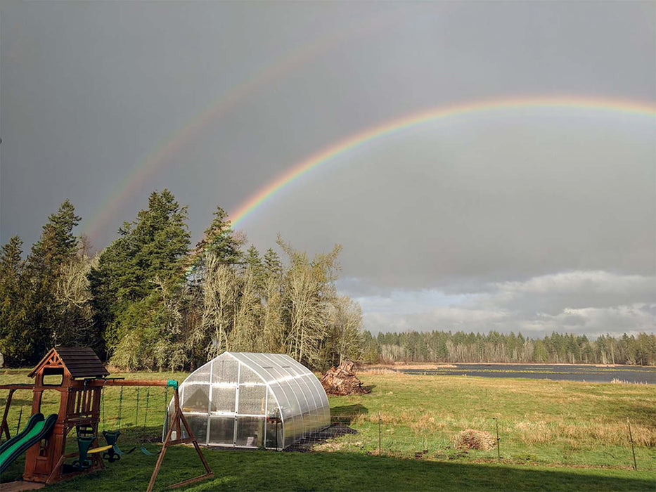 Exaco Hoklartherm Riga XL 6 Greenhouse in an open field beneath a double rainbow, blending natural beauty with durable year-round gardening design.